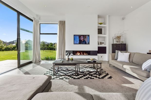 Bright contemporary living room with expansive floor-to-ceiling sliding glass doors opening onto a green lawn and landscaped garden. The room features a large neutral sectional sofa, upholstered ottoman, patterned geometric rug, glass-topped coffee table, built-in shelving, and a wall-mounted television above a linear fireplace. White walls, recessed lighting, and abundant natural light create an airy, refined interior.