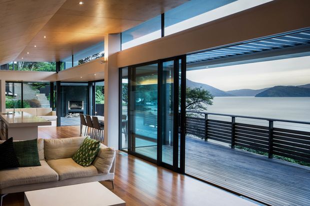 A contemporary open-plan living area with timber flooring and a warm timber ceiling, furnished with a large neutral sofa and patterned cushions. Full-height glazed sliding doors open onto a broad timber deck with dark horizontal balustrading, overlooking a calm lake and surrounding hills. The interior also includes a kitchen island, dining area, and a fireplace visible deeper in the space. Strong indoor-outdoor connection and expansive glazing define the composition.