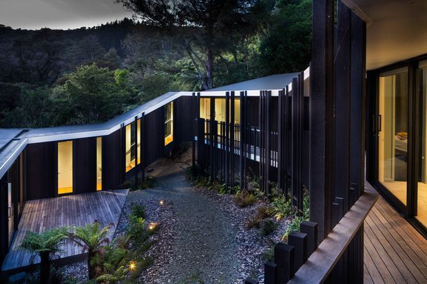 A dramatic dusk view of a low, modern multi-wing residence set into a densely vegetated, steep bushland site. The dark vertical-clad buildings have angular low-pitched rooflines, extensive warm-lit glazing, timber decking and screened walkways arranged around a landscaped gravel courtyard with native planting.