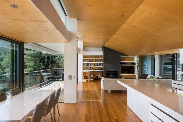 Open-plan contemporary living, dining, and kitchen interior with extensive floor-to-ceiling glazing opening to a balcony or terrace. The space features a warm timber-lined ceiling, polished timber flooring, a large white kitchen island with integrated cabinetry, a dining table, built-in timber shelving, and a dark feature wall with fireplace and media joinery. The room is bright and connected to surrounding greenery.