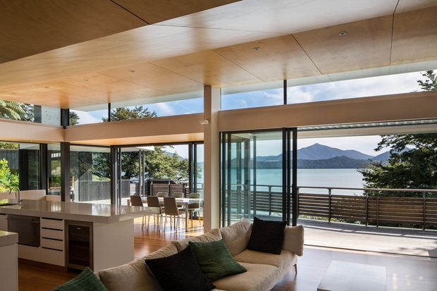 Open-plan contemporary living and kitchen interior with extensive floor-to-ceiling glazing and sliding glass doors opening onto a deck. The space has a timber-lined ceiling, light timber flooring, a pale kitchen island with dark cabinetry, and a large neutral sofa. Through the glazing is a calm lake and mountain landscape, while a timber-and-metal deck balustrade frames the outdoor view.
