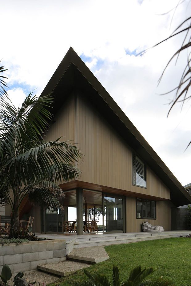 Exterior view of a contemporary single-storey dwelling with a steep triangular gable roof, dark roof trim, vertically ribbed beige-brown wall cladding, and extensive ground-level glazing opening onto a raised concrete terrace. Tropical planting, a lawn, concrete steps, and low block retaining walls surround the house.
