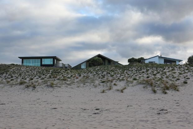 Wide exterior view of several low-profile contemporary beach houses set behind a sandy dune, with large glazing, simple geometric roof forms, and sparse coastal vegetation under an overcast sky.