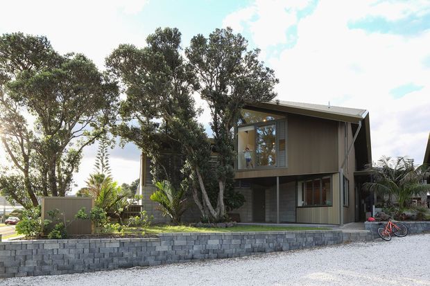 Exterior view of a contemporary two-storey residence set behind a low stacked-block retaining wall and landscaped garden. The house has dark vertical cladding, extensive glazing, a projecting upper level, and a simple sloping roof with deep eaves. A gravel driveway and bicycle are visible in the foreground, with mature trees and tropical planting surrounding the building.