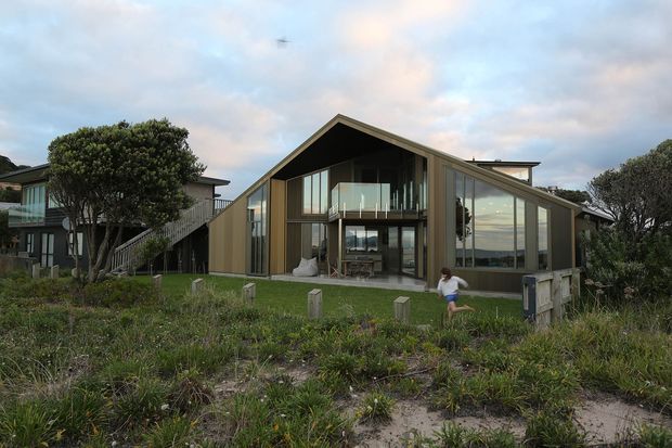 Exterior view of a contemporary coastal residence with a dramatic steep gable roof, extensive floor-to-ceiling glazing, and vertical timber-toned cladding. The home sits behind dune vegetation, with a raised adjoining structure and outdoor living areas visible through the glass. The central glazed gable exposes a double-height interior volume and balcony, while the muted natural palette integrates with the seaside setting.
