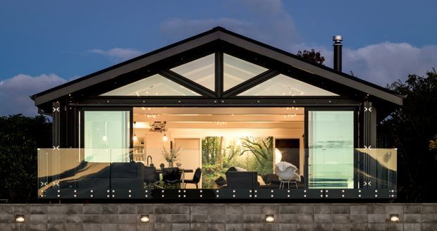 Exterior twilight view of a contemporary single-storey pavilion-style home with a prominent glazed gable facade. The open living area is visible through full-height glazing, with a balcony or terrace enclosed by transparent glass balustrades. The building features dark structural framing, a pitched gable roof, warm interior lighting, and a low masonry retaining wall with integrated lights.