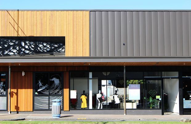 Street-level view of a contemporary commercial building facade with warm vertical timber cladding, dark grey vertically ribbed metal cladding, large shopfront glazing, and a decorative black breeze-block screen above the entrance. Retail interiors and mannequins are visible through the windows.