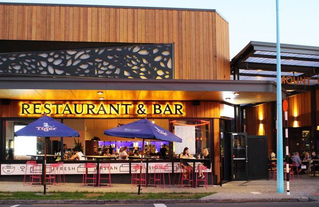 Street-facing exterior of a contemporary restaurant and bar, with warm vertical timber cladding, a dark perforated decorative facade panel, illuminated signage, outdoor bar seating beneath branded umbrellas, and a covered entry terrace. The image primarily shows a commercial hospitality frontage in an urban streetscape.
