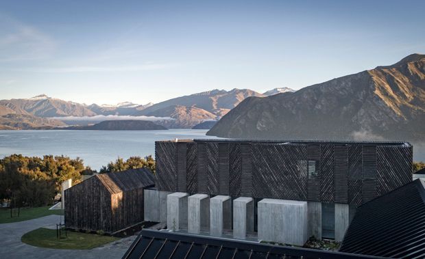 Exterior view of a contemporary dark-clad building complex set beside a lake, with a rugged mountain range in the background. The main structure has a large rectangular form, vertically varied dark timber-like cladding, narrow windows, and a series of pale concrete volumes at its base. Smaller steep-roofed dark-clad buildings surround it.