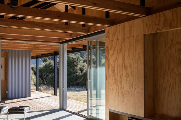 Interior view of a timber-lined contemporary space opening onto a timber deck through large sliding glass doors. Exposed timber beams and rafters span the ceiling, while plywood wall panelling and aluminium-framed glazing create a warm, minimal architectural character. A small seating area is visible in the foreground, with a bushy landscape beyond the open connection to the exterior.
