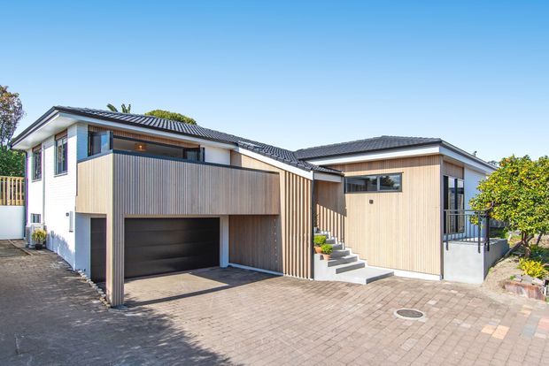 Exterior view of a contemporary multi-level residential home with two connected pitched roof volumes. The facade combines light vertical timber cladding, white rendered walls, dark grey tiled roofing, large windows, and an integrated dark garage beneath a projecting upper level. A paved driveway and front steps lead to the entry, with established planting around the edges.