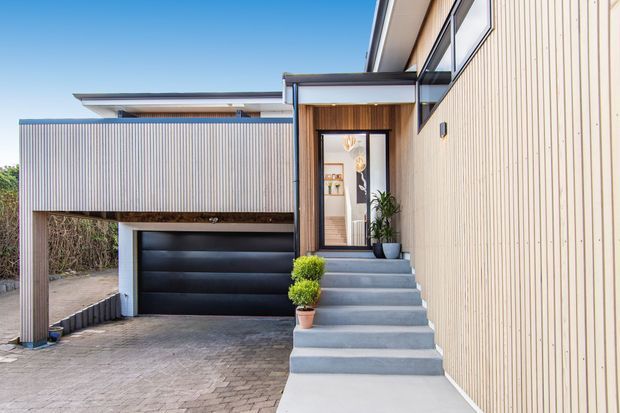Contemporary multi-level residential facade with a strong vertical timber-clad entry wall, contrasting pale corrugated cladding, a black sectional garage door, and a broad concrete stair leading to a glazed front door. The home has crisp geometric lines, flat roof forms, timber soffit detailing, and a paved driveway.