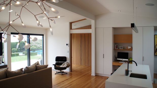 Bright contemporary open-plan living area with a compact kitchen in the foreground. The room features warm timber flooring, white walls and built-in cabinetry, a brown upholstered sofa, a sculptural branching pendant light, and full-height sliding glazing opening toward a landscaped garden and pool. A timber slatted feature screen and a small built-in study nook add architectural detail.