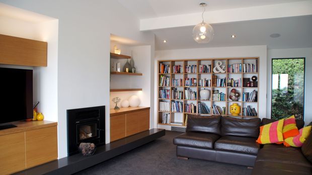 A warm, contemporary living room with a dark leather sectional sofa, built-in timber cabinetry and shelving, a black fireplace surround, television unit, and a large floor-to-ceiling bookcase filled with books. Recessed ceiling lights and a spherical pendant provide illumination, while a colourful striped cushion adds a small accent. A window brings in greenery from outside.