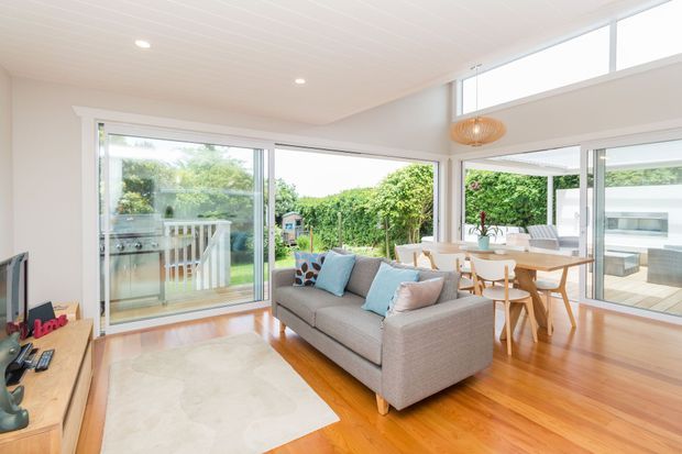 Bright open-plan living and dining room with extensive floor-to-ceiling glazing and sliding doors opening onto a deck and leafy garden. The room features warm timber flooring, white painted walls and ceiling, a grey upholstered sofa, light dining furniture, and a woven pendant light.