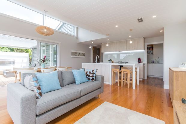 Bright open-plan contemporary living and kitchen area with a large grey sectional sofa in the foreground, light blue and patterned cushions, warm timber flooring, white cabinetry, a kitchen island with timber stools, dining area, and extensive glazing opening toward an outdoor space. The room has a light, airy feel with recessed and pendant lighting.