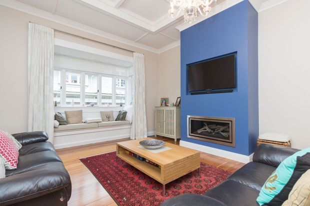 Bright, refined living room with a bold blue feature wall containing a wall-mounted television and built-in fireplace. The room features black leather sofas, a large patterned red rug, a timber coffee table, a window seat with layered cushions, white curtains, decorative ceiling moulding, and a crystal chandelier.