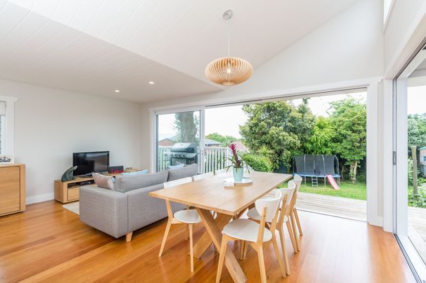 Bright open-plan living and dining area with warm timber flooring, white walls and ceiling, a light grey sectional sofa, timber dining table with matching chairs, and large sliding glass doors opening to a leafy garden. A woven pendant light and extensive glazing create an airy, family-oriented interior.