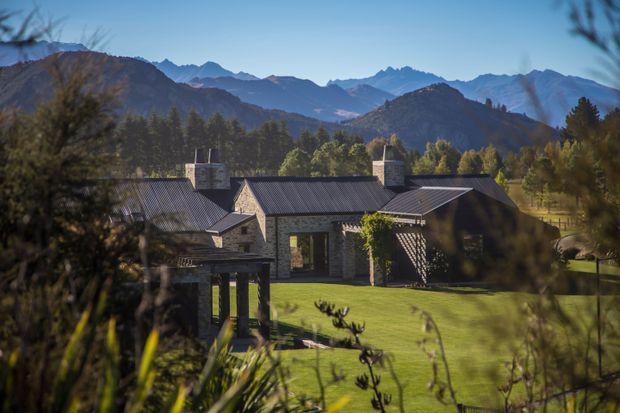 A low-slung stone and dark metal-roofed house set in a landscaped rural or alpine setting, viewed through foreground vegetation. The building has several connected pitched-roof volumes, chimneys, timber-framed openings and a covered outdoor structure, with dramatic mountain ranges and mature trees behind it.