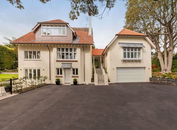 Front exterior view of a substantial cream-rendered residential house with a terracotta tiled, multi-gabled roof. The facade features white-framed multi-pane windows, a central covered entry reached by steps, and a connected garage wing with a double garage door. A broad dark paved driveway, mature trees, garden beds, and a black metal gate frame the property.