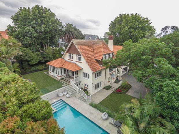 Elevated aerial view of a substantial traditional two-storey house set within a mature landscaped garden. The home has cream rendered walls, red terracotta tiled pitched roofs, white-framed windows, verandah-like covered areas, and a paved outdoor terrace beside a rectangular swimming pool. Dense established trees and palms surround the property.