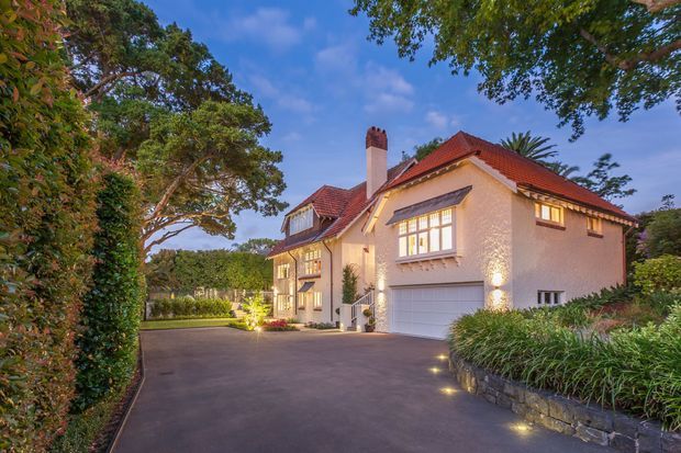 Exterior view of a substantial traditional-style rendered house at dusk, featuring red tiled pitched roofs, dormer windows, a prominent brick chimney, illuminated multi-pane windows, and an integrated double garage. The broad paved driveway is bordered by landscaped garden beds, stone retaining walls, and mature trees.