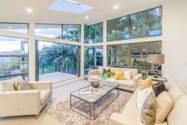 Bright, spacious contemporary living room with large floor-to-ceiling windows and glazed doors opening to a timber deck and leafy garden. The room features cream upholstered sofas and armchairs, a glass-topped metal coffee table, a large textured rug, recessed lighting, and a roof skylight. Reflections in the glazing show the interior extending around the room.