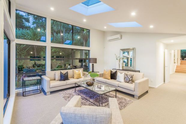 Bright contemporary living room with two cream sofas, a matching armchair, glass coffee table, patterned area rug, and neutral finishes. Floor-to-ceiling glazing overlooks lush greenery, while multiple skylights bring additional daylight into the room. A hallway and steps are visible beyond the living area.