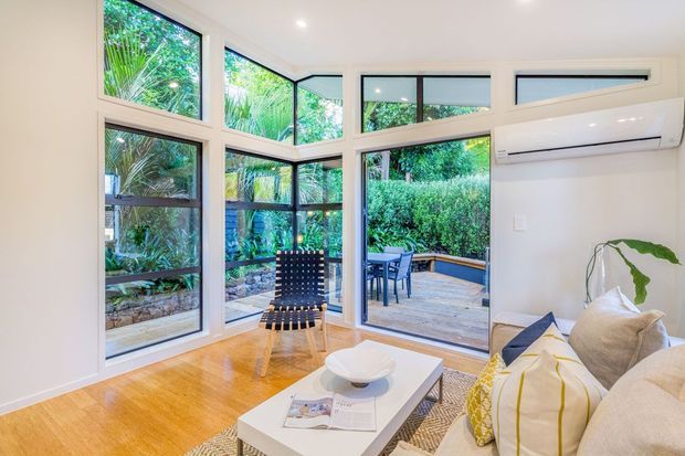 Bright contemporary living room with polished timber flooring, large corner windows and glazed doors opening to a timber outdoor deck and lush garden. The room includes a cream sectional sofa, white coffee table, patterned cushions, and a wall-mounted split-system air conditioner.