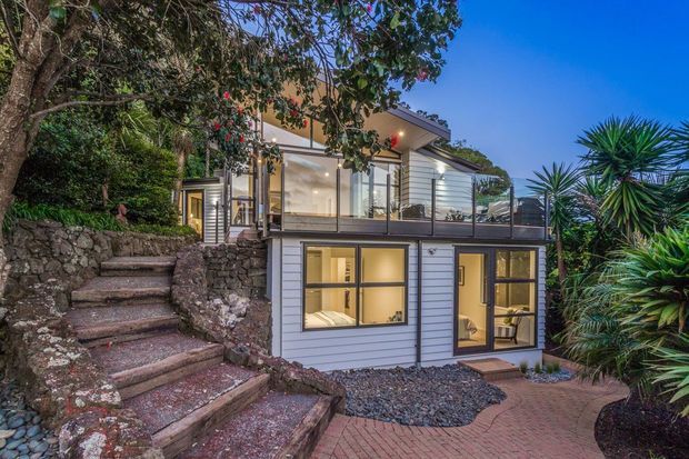 A contemporary multi-level residential house built into a landscaped, rocky sloping site. The exterior features pale horizontal weatherboard cladding, extensive glazing, a glass-balustraded upper terrace, warm interior lighting, and a paved brick pathway with stone steps and tropical-style planting.