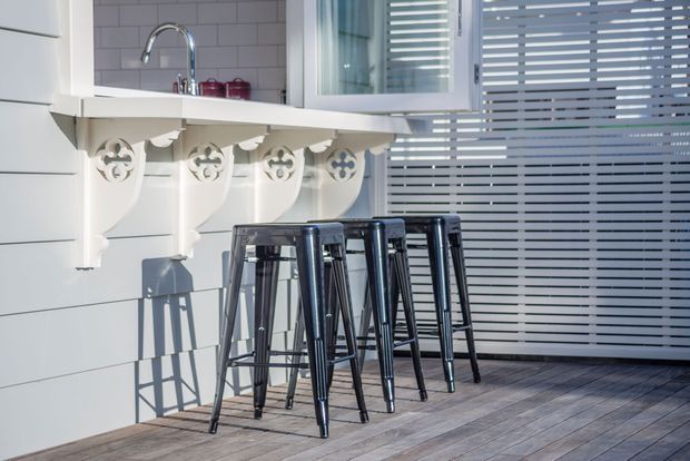A bright, compact kitchen or entertaining counter with a white-painted wall-mounted bench, decorative corbel brackets, and a chrome gooseneck tap over a tiled backsplash. Several black metal bar stools line the counter. Horizontal blinds cover the glazing on the right, and grey-toned timber-look flooring extends across the space. The design combines crisp white surfaces with traditional decorative detailing and industrial-style seating.