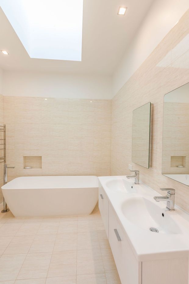 Bright contemporary bathroom featuring a freestanding white bathtub, double vanity with two basins and chrome taps, large wall mirrors, pale beige stone-look wall and floor tiles, recessed lighting, and a large rectangular skylight.