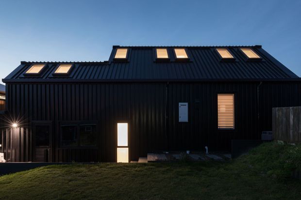 Exterior view of a dark, vertically ribbed metal-clad building at dusk. The steep pitched roof has multiple skylights, and the facade includes a small illuminated entry door, a louvered window, and dark-framed openings. A grassy foreground and timber boundary fencing surround the structure.
