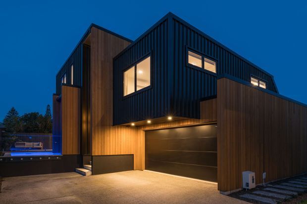 A contemporary multi-level residential exterior photographed at dusk. The home combines dark vertically ribbed cladding with warm timber-clad wall sections, illuminated windows, a projecting upper volume, and an integrated double garage. A paved driveway and exterior lighting are visible.