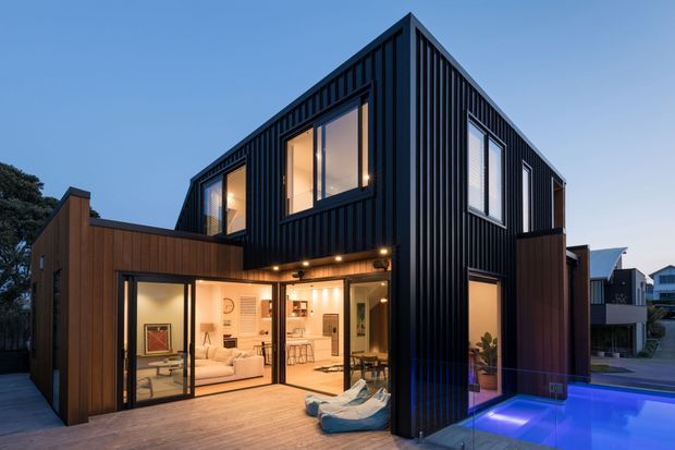 Twilight exterior view of a contemporary multi-level home with a sharply pitched gable-like roof form, dark vertically ribbed cladding, warm timber-clad lower volumes, extensive glazing, and a small illuminated swimming pool beside a timber deck. Warm interior lighting and visible living areas create an inviting contrast with the dark facade.