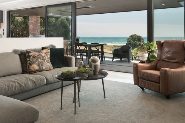 A contemporary coastal living room with a large grey sectional sofa, brown leather armchair, nesting round coffee tables, and soft neutral furnishings. Full-height sliding glass doors open to an outdoor dining terrace with unobstructed ocean views. The room has a polished concrete floor, white walls, dark-framed glazing, and abundant natural light.