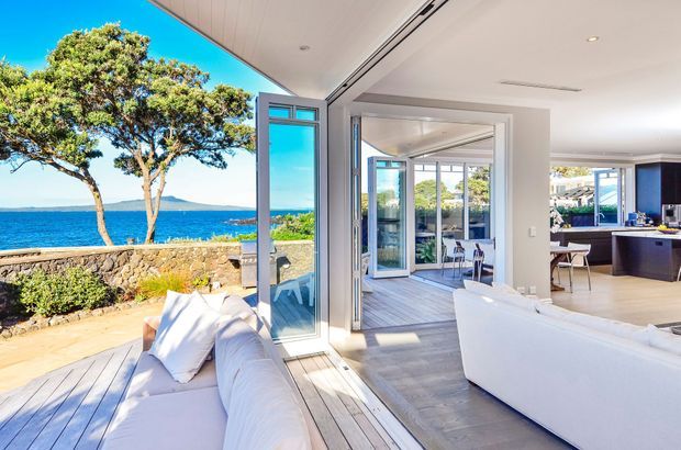 Bright coastal home interior opening directly onto a timber deck, with expansive folding glass doors, a white sofa in the foreground, and an open-plan living and kitchen area beyond. A stone boundary wall, mature trees, blue water and distant shoreline are visible outside. The space has a clean, contemporary design with pale finishes and strong indoor-outdoor connection.