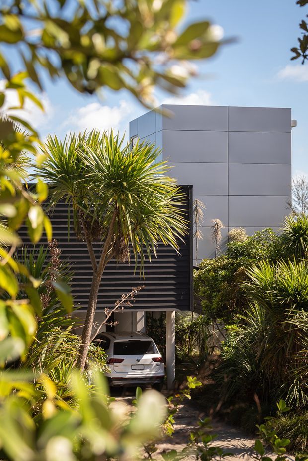 Exterior view of a contemporary multi-level house set within dense landscaped planting. The building features a boxy form clad in large pale grey panels, a dark horizontal-slatted screen, and a sheltered carport with a vehicle beneath. Tropical and subtropical foliage frames the sunny residential scene.
