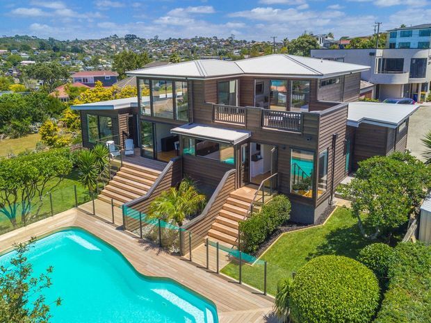 A multi-level contemporary residential home on a landscaped suburban site, viewed from above. The dark horizontal timber-clad exterior has extensive glazing, several pitched metal roof sections, balconies and decks, and broad timber stairs leading down to a turquoise swimming pool enclosed by frameless-style glass pool fencing. Lush gardens and neighbouring homes are visible in the background.