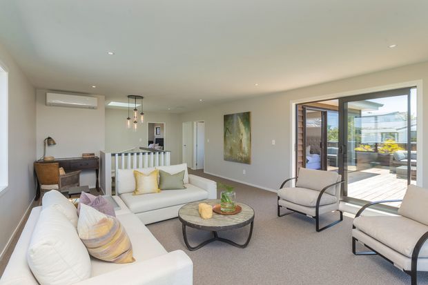 Bright contemporary living room with white sofas and armchairs arranged around a round coffee table, a compact desk area, neutral carpet, recessed ceiling lights, and a large glazed opening leading to an outdoor deck or courtyard. The room has soft neutral finishes with yellow and pastel accent cushions, plus a partial view of an adjacent kitchen or stair area.