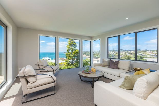 Bright contemporary living room with expansive corner windows overlooking a coastal residential landscape and ocean. The room features pale upholstered armchairs and sofas, a round coffee table, neutral carpeting, recessed ceiling lights, and a calm, airy palette.
