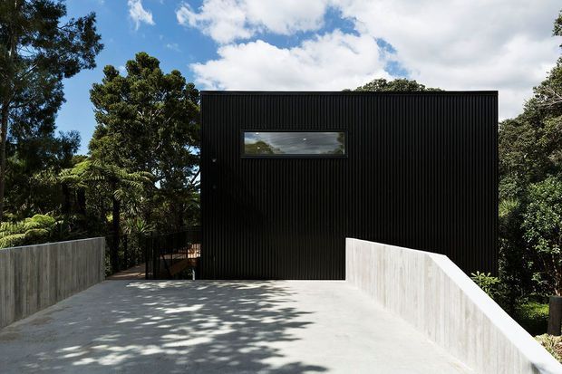 Exterior view of a minimalist black-clad building set within mature greenery. The rectangular facade has vertical ribbed cladding, a narrow horizontal window, and a flat roof, opening onto a broad pale concrete driveway or terrace enclosed by sculptural concrete retaining walls.