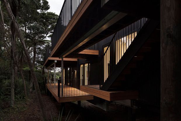 Dark, contemporary elevated timber-and-metal building integrated into dense bushland. The image focuses on projecting timber decks, black vertical balustrades and cladding, exposed structural beams, and an external stair connecting levels. Warm interior lighting glows through the glazing, creating a dramatic forest setting.