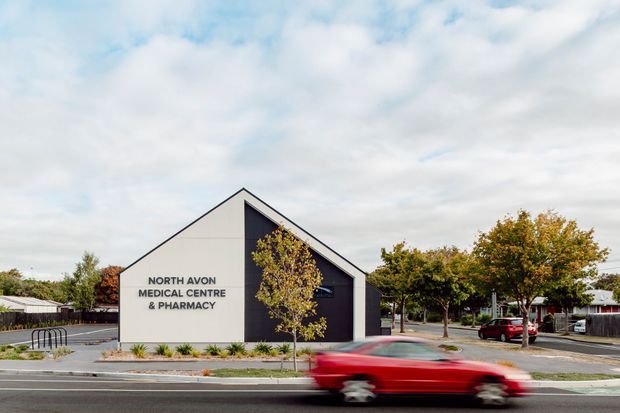 Street-level exterior photograph of a small contemporary medical centre and pharmacy in a suburban commercial setting. The building has a simple steep gable form, pale rendered side wall, dark vertical front facade, prominent signage, landscaped frontage, parking, and a blurred red car passing in the foreground.