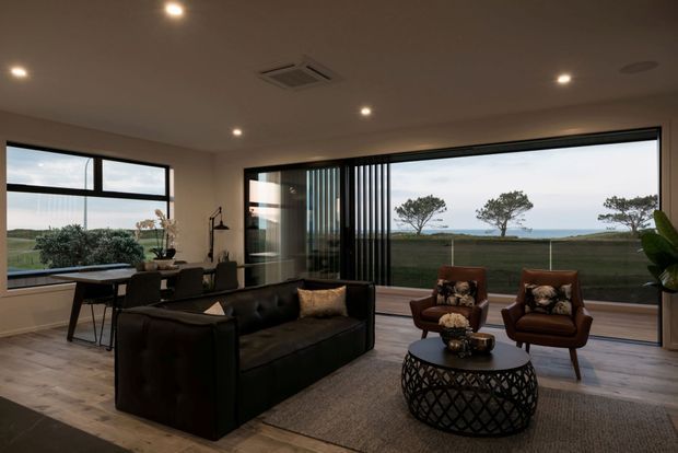 Contemporary living room with expansive floor-to-ceiling glazing overlooking a coastal landscape. The room features a black leather sofa, two brown leather lounge chairs, a round lattice coffee table, neutral rug, timber-look flooring, recessed ceiling lights, and dark-framed sliding doors with vertical screening.
