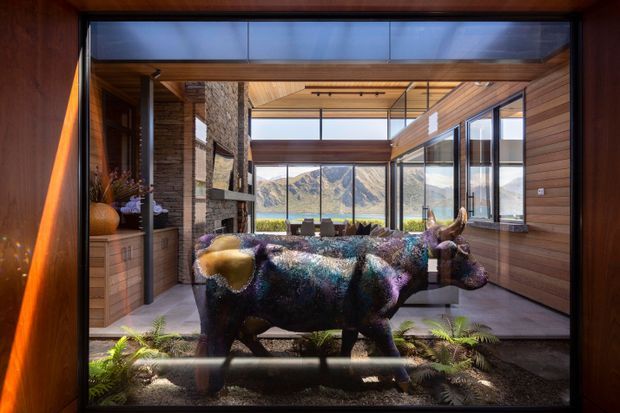 A dramatic contemporary interior with extensive timber wall and ceiling lining, dark stone masonry, and floor-to-ceiling glazing overlooking rugged mountains and water. A large sculptural bull artwork occupies the foreground, with built-in timber cabinetry, a fireplace or stone feature wall, and a seating area beyond. The space has strong indoor-outdoor visual connections and abundant natural light.