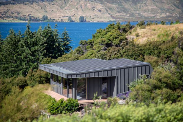 A compact contemporary single-storey house with dark vertical metal cladding and a low-pitched metal roof, set among native vegetation on a steep lakeside or waterfront site. Large glazed openings and a covered deck face the vivid blue water and rugged hills beyond.