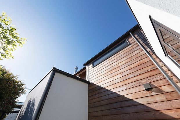 Upward-looking exterior view of a contemporary residential building, showing warm horizontal timber wall cladding, white rendered wall surfaces, dark metal roof and guttering, and angular roof forms against a clear blue sky.