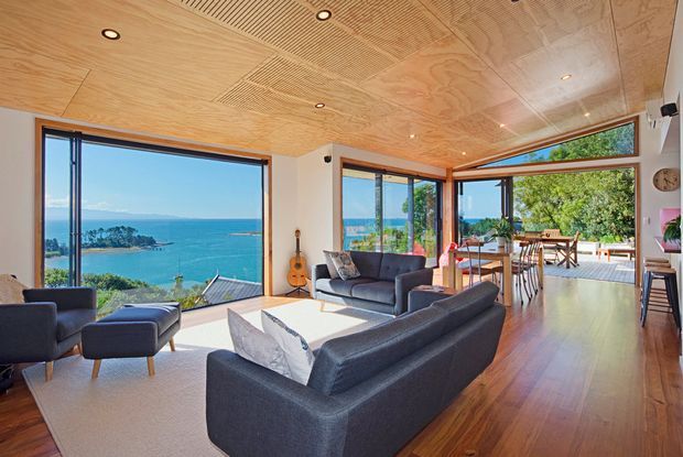 Bright contemporary open-plan living room with expansive timber-framed glazing overlooking a turquoise coastal bay. The room features dark upholstered sofas and ottomans, a warm timber floor, a plywood-lined ceiling with recessed downlights and patterned acoustic panels, and an adjoining dining area opening onto a timber deck.