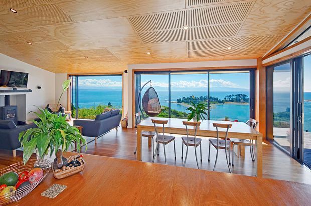 A bright open-plan living and dining area with expansive floor-to-ceiling glazing overlooking a coastal bay. The room features warm timber flooring, a timber-lined ceiling with integrated linear ventilation grilles and recessed lighting, dark timber-framed glazing, a dining table with six chairs, a grey sofa, fireplace, indoor plants, and a kitchen island or countertop in the foreground.